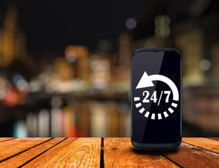 24 hours service icon on modern smart phone screen on wooden table over blur light city tower background, Full time service concept - 119541730
