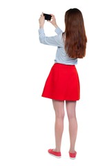 back view of standing young beautiful woman using a mobile phone. Long-haired brunette in red skirt holding phone horizontally.