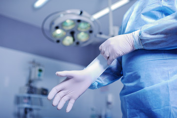 Surgeon putting on sterile rubber gloves to complicated surgical operation on the background lights. Surgery - obrazy, fototapety, plakaty