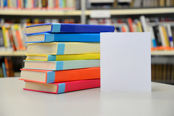 Close up of colorful books on the table. Blank white card beside the books. Bookshelves in the background.