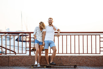 Young beautiful couple walking at seaside, smiling, skateboarding.