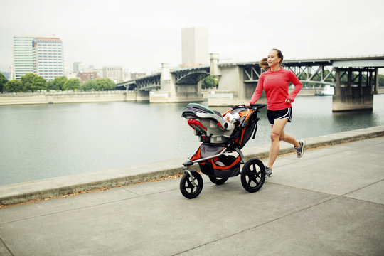 Woman jogging with baby stroller on footpath by river against bridge