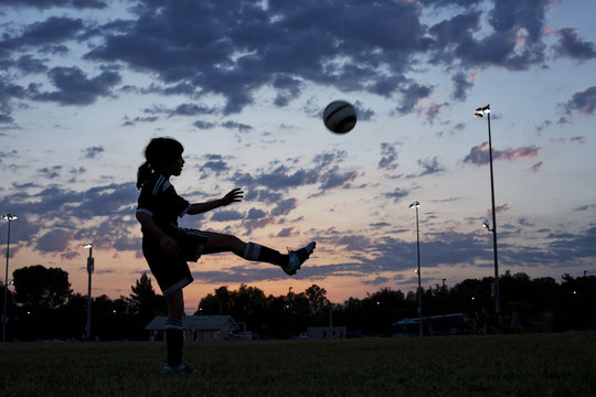 Side view of girl kicking soccer ball at field against sky during sunset