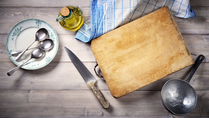 Cutting board, knife, oil cruet, strainer, dinner plate with spoons, tea cloth on old wooden background