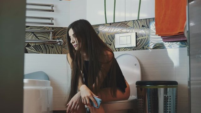 Brunette young girl sitting on toilet with smartphone in hands. Bathroom. Apartment