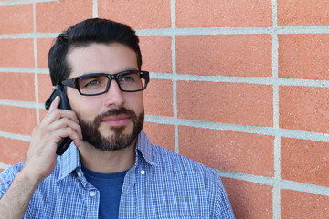 Young bearded man with glasses talking on mobile phone isolated on red brick wall modern background