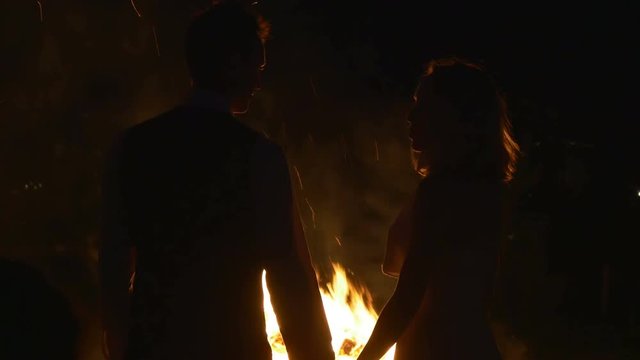 Silhouettes of man and woman in love. They stand near bonfire holding their hands