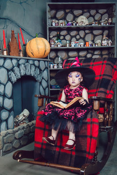 Girl in witch costume and make-up on her face sitting in a chair with  book in his hands in halloween decorations