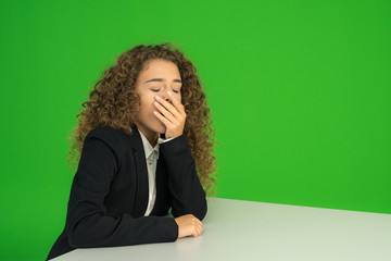 The young girl sit at the table and yawn on the green background