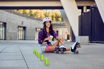 Fototapeta na wymiar Young woman take a rest after riding roller skates and drinking coffee or tea sitting on the pavement on street. Happy girl enjoying roller skating rollerblading in urban park.