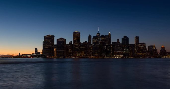 New York City Lower Manhattan skyscrapers between sunset, dusk and nightfall. Time lapse cityscape view of the Financial District lights and East River with passing boats