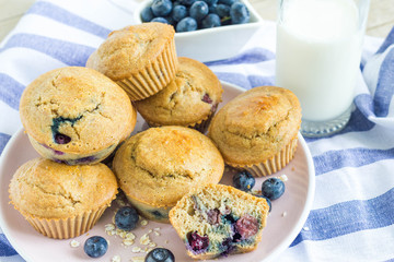 Bunch of oatmeal blueberry muffins with a glass of milk and a bowl of blueberry in the back placed on a blue tablecloth.