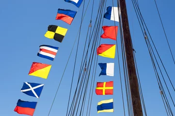 Wallpaper murals Sailing Colorful nautical sailing flags flying in the wind from the lines of a sailboat mast backlit in bright blue sky by the sun