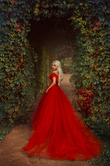Beautiful blonde girl with long hair. She is in a luxurious red dress. Queen walks near the castle. Vampire passion, gothic story. Smoky eyes and red lips.  