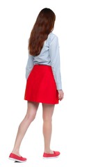 back view of walking woman. Long-haired brunette in red skirt moves slowly.
