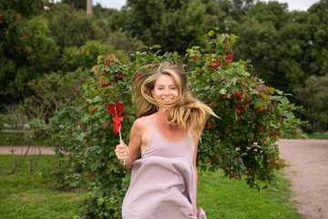 The young girl with a fair long hair dressed in a pink dress holds in hand lollipop in shape heart