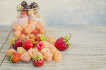 Multicolored raspberries in a glass jar with strawberries on background