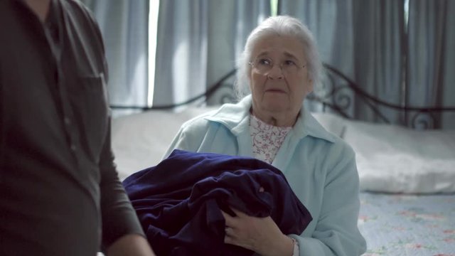 A senior woman on her bed suffers elder abuse when an abusive caretaker throws clothes at her. Shot in 4K UHD.