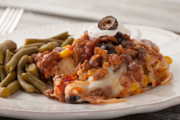 Fiesta bake Mexican lasagna with corn tortillas, pinto beans, olives, corn, tomatoes, and sour cream served with green beans macro shot on a weathered barn wood table