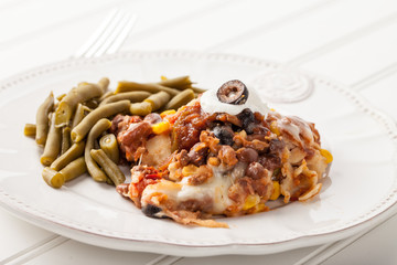 Fiesta bake Mexican lasagna with corn tortillas, pinto beans, olives, corn, tomatoes, and sour cream served with green beans side view on white wood