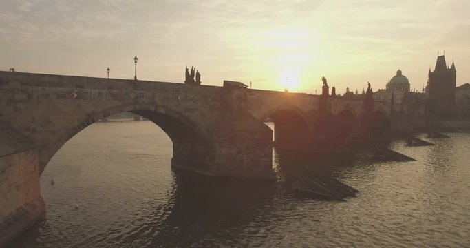 Aerial camera flies fast along the Charles Bridge in Prague as the sun rises behind it. 