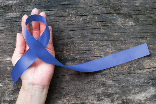 Dark blue ribbon on woman hand, old aged wood background: Satin fabric silk ribbon symbolic concept raising awareness concerns help campaign on people living with Colorectal cancer illness