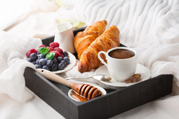 Morning breakfast in bed with cup of coffee, croissants, fresh berries and honey on wooden tray, selective focus