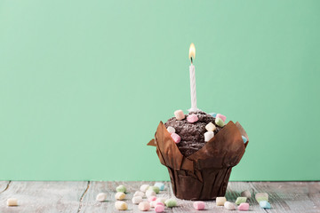 Delicious birthday chocolate muffin with colorful marshmallows and a single candle on a green mint background, copy space