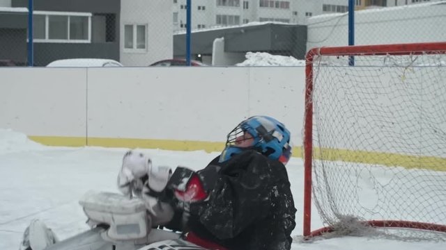 Slow motion of masked hockey goaltender player trying to catch puck in catchers during outside drill, then missing it and falling down on ice