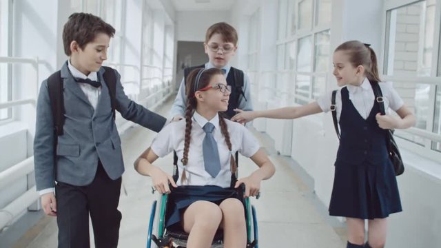 Handheld dolly-like shot of schoolchildren with backpacks running along school hallway towards handicapped schoolgirl in uniform and glasses riding alone in wheelchair, then chatting and laughing