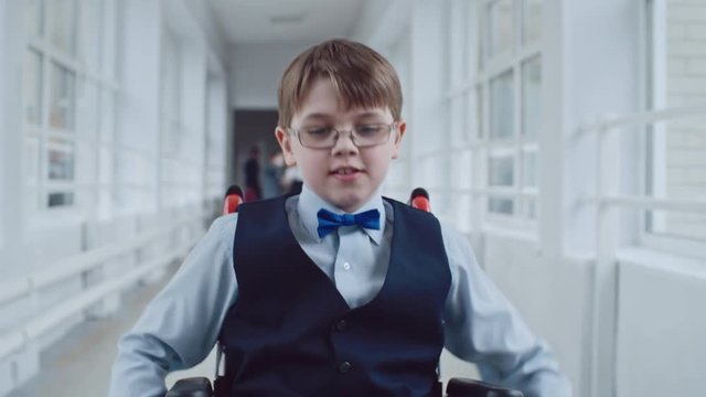 Handheld dolly-like shot of concentrated handicapped schoolboy in wheelchair wearing uniform and glasses riding along school hallway during recess