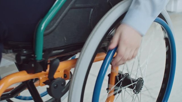 Closeup of disabled or recovering little boys hand turning wheels on his wheelchair and riding along hallway 