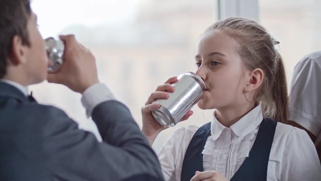 Camera moving from chatting schoolboy and schoolgirl in uniforms drinking soda from tin cans to smiling schoolchildren in glasses listening music on mobile phone 