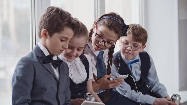 Medium shot of schoolboy in uniform making silly faces to tablet, then showing something to smiling schoolgirl holding soda and classmates in glasses listening to music and dancing