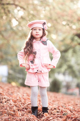 Stylish kid girl 5-6 year old wearing trendy winter jacket, shorts and hat standing in fallen leaves outdoors. Looking at camera. Autumn season. Posing child girl.