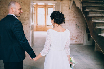 Stylish bride and groom holding hands in a room with white brick. Loft