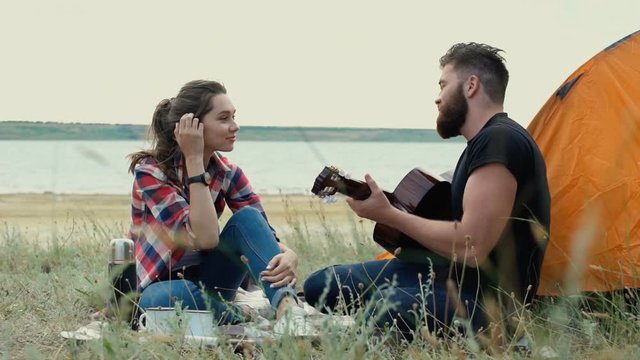 Young handsome bearded man playing guitar and having drink with his girlfriend