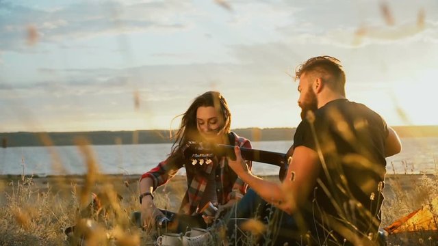 Young bearded man playing guitar for his girlfriend while she is pouring cup of tea from thermos