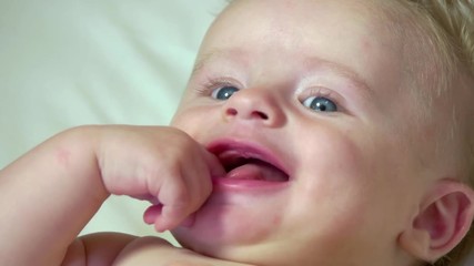 Portrait of child having fun in cradle at home, happy cute little boy laughing and thumb sucking in bed. Male infant with blue eyes in baby crib. Close-up of face, slow motion