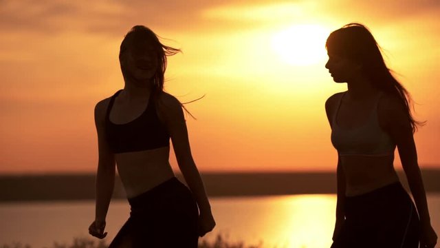 Silhouettes of two beautiful girls dancing zumba in field at sunrise. Slow motion.