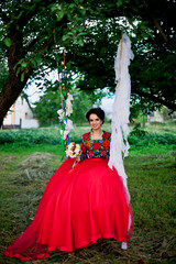 Fototapeta premium Charming bride in amazing red dress is sitting on swing