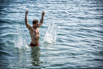 Handsome, hot young bodybuilder in the sea, splashing water up, showing his muscular torso and arms