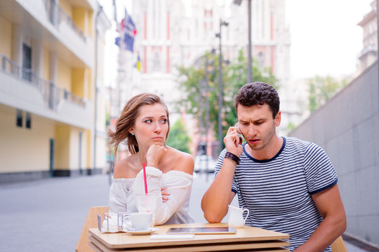 Young couple in sidewalk cafe. Attractive woman is angy with her boyfriend, who talking by phone.