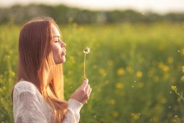 Young spring fashion woman blowing dandelion in spring garden. Springtime. Trendy girl at sunset in...