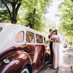 Happy bride and groom hugging and posing near old retro car limousine before wedding ceremony