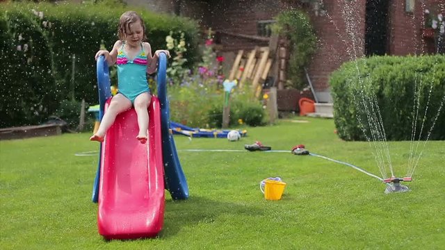 Smiling little girl having fun with garden sprinkler and children's slide in the backyard, slow motion
