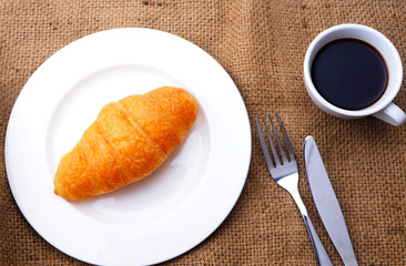 Top view of breakfast with croissants and a cup of black coffee on a sackcloth background.