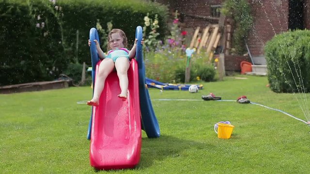 Smiling little girl having fun with garden sprinkler and children's slide in the backyard, slow motion
