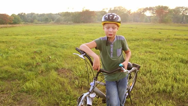 Child in cycle helmet talking cheerfully to camera near his bike. Little 9 years old boy having conversation with somebody outside over green nature background on summer day. Real time video footage.