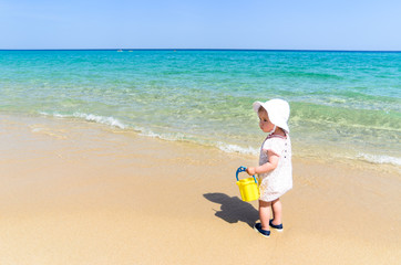 Adorable little girl in beautiful swimsuit and white hat holding kid bucket and having fun at exotic white sandy beach during vacation over the background of turquoise sea and blue sky, copy space